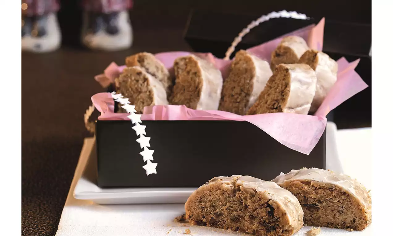 Alpenbrot mit braunen Stückchen und heller Zuckerglasur, präsentiert in einer schwarzen Schachtel mit rosa Papier auf weißem Teller