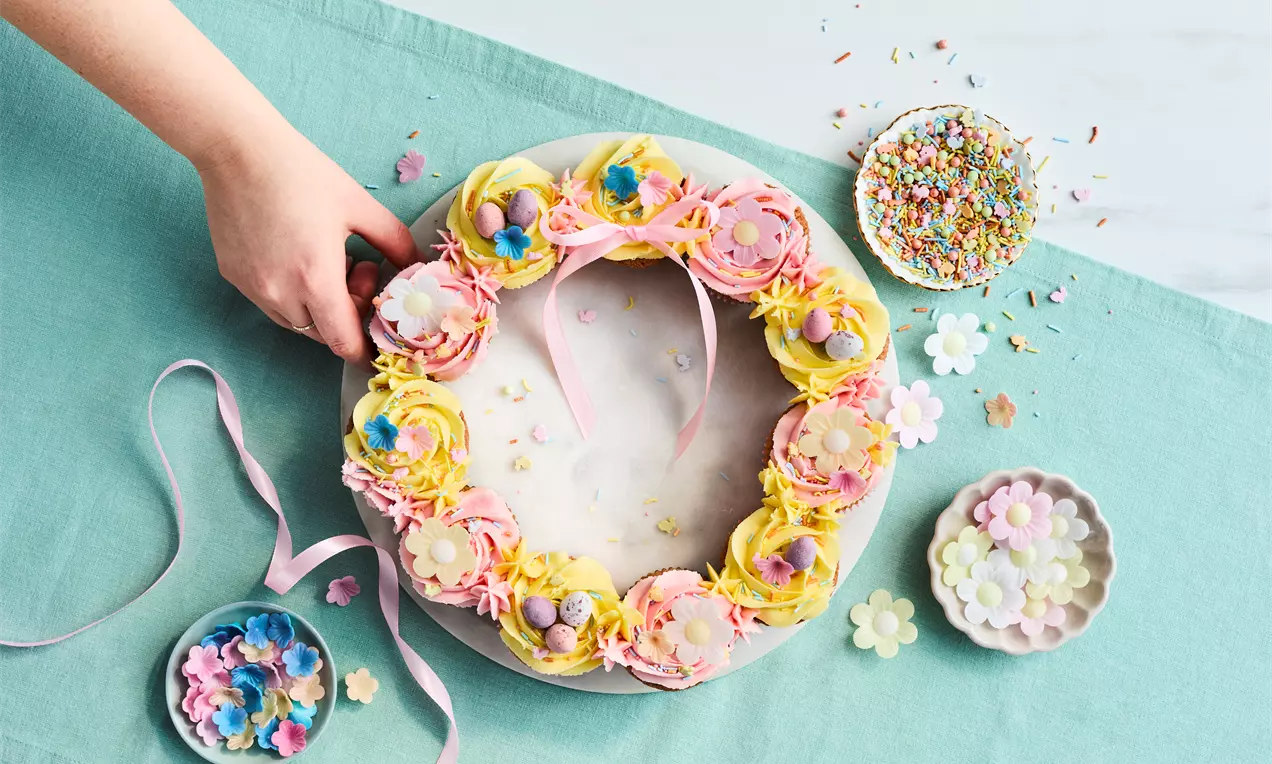A wreath of pastel cupcakes with yellow and pink swirls, flower decorations, and ribbon, arranged on a white plate with scattered sprinkles