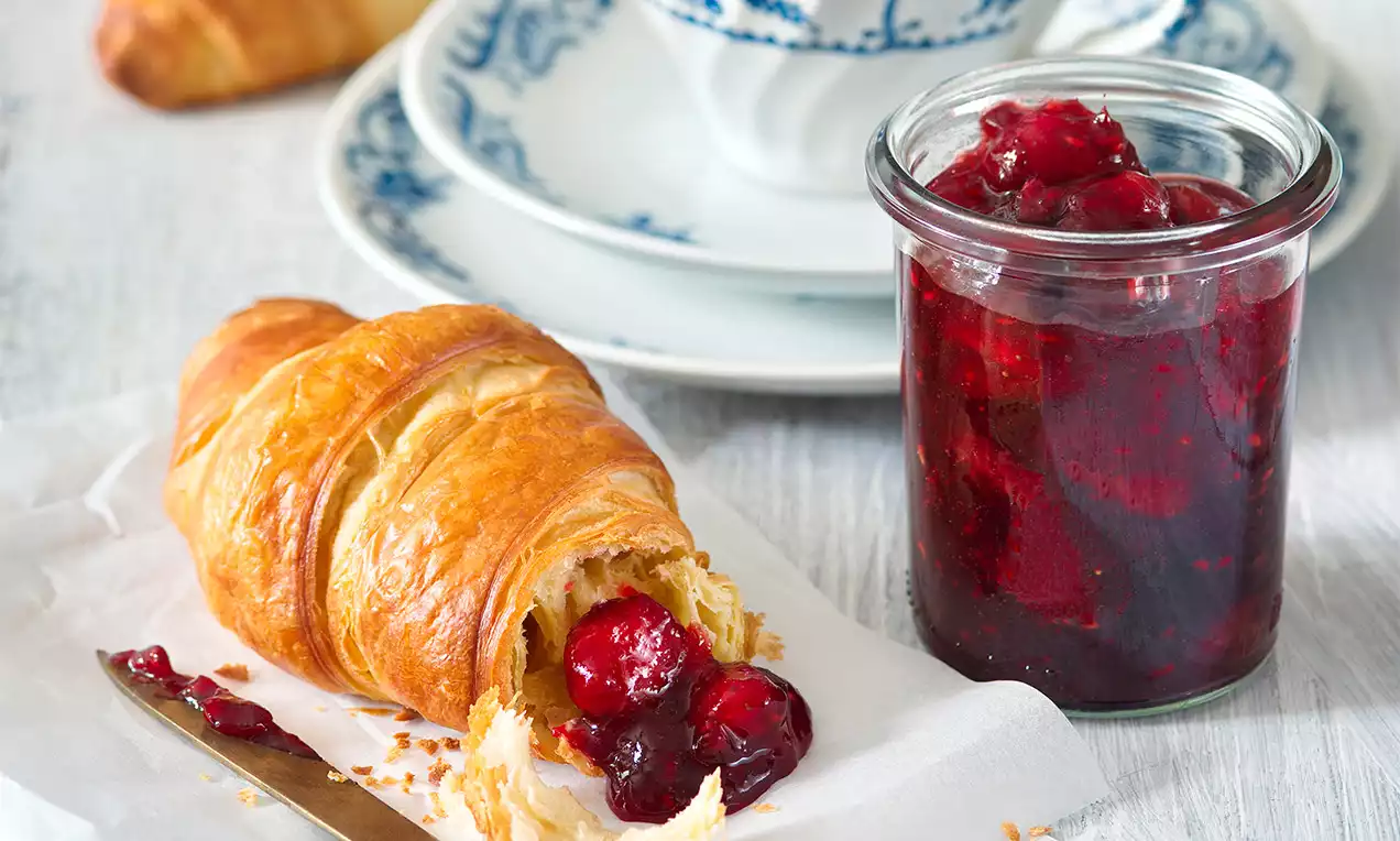 Zlatistý croissant s jemne nakrčeným povrchom plnený lesklým višňovým džemom vedľa skleného pohára s rubínovo červenou marmeládou na bielom papieri