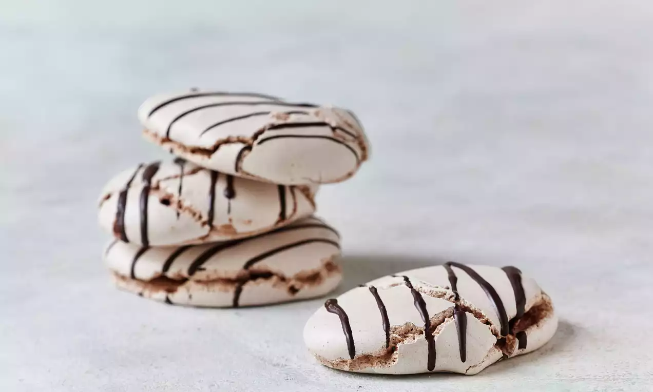Sprøde, lysebrune marengs med spinkle chokoladestriber, ujævne kanter og luftige indre stablet og liggende på en lys flade