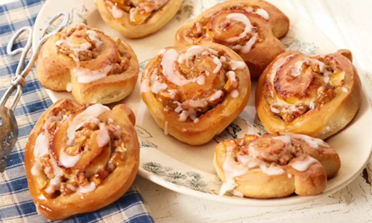 Goudbruine kaneelbroodjes met glanzende glazuurspiraaltjes en stukjes appel en nootjes, gerangschikt op een lichtgekleurd bord