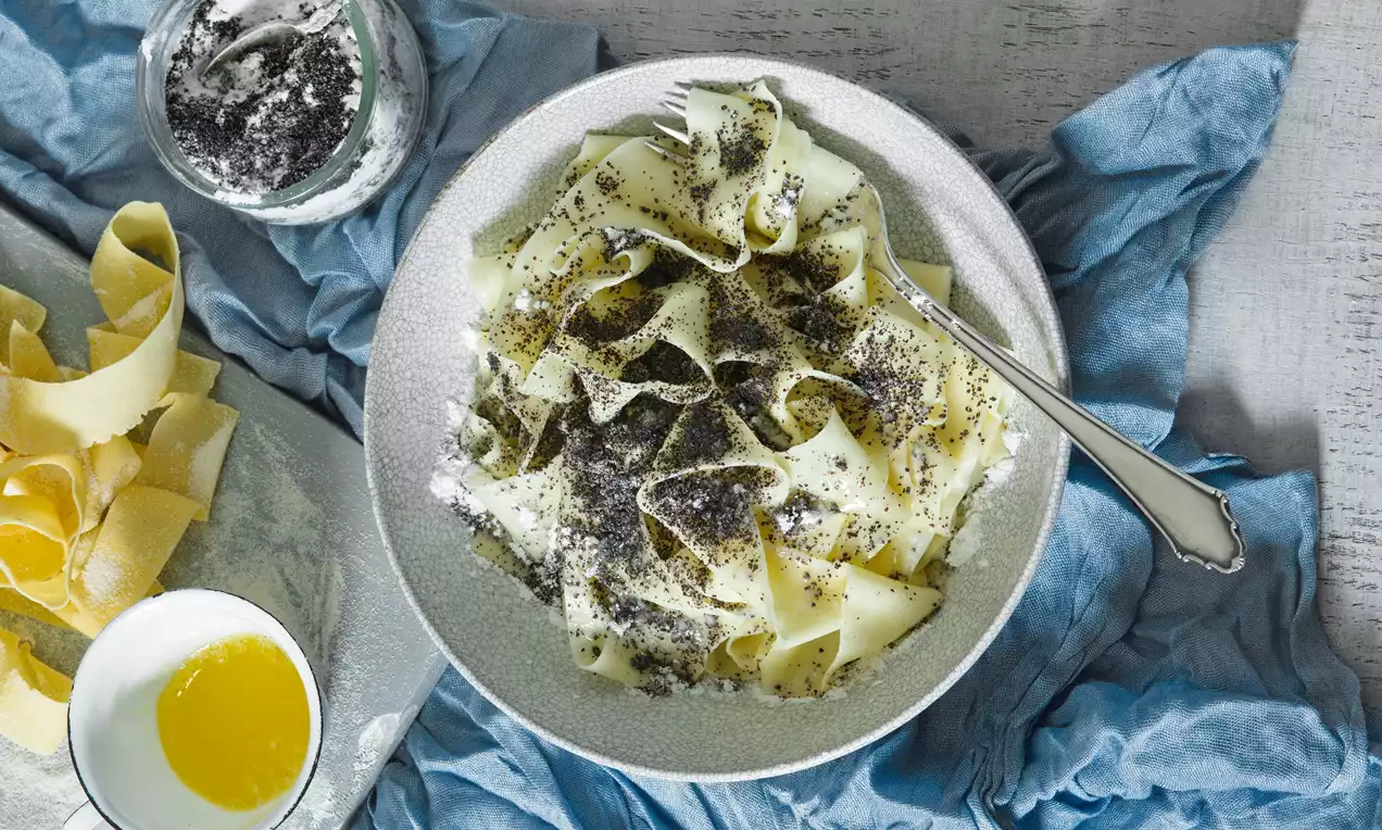 Široké světle žluté nudle na keramickém talíři, bohatě posypané černým mletým mákem a bílým cukrem, vedle rozpuštěné máslo a modrá látka