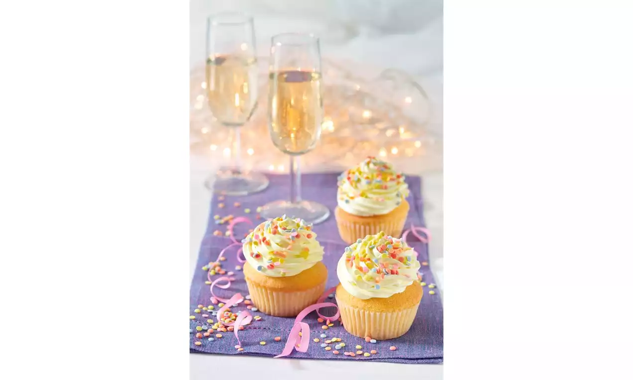 Silvestrovské cupcakes zlatisto žlté s nadýchaným krémom ozdobené farebnými cukrovinkami na fialovej servítke vedľa pohárov šampanského