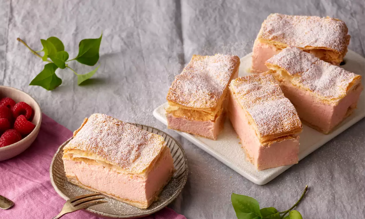 Himbeer-Cremeschnitten mit rosa Füllung zwischen goldbraunen Blätterteigschichten, bestäubt mit Puderzucker, angerichtet auf Tellern und Platte