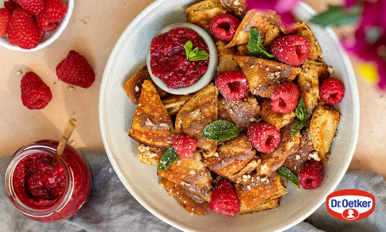 Malinový džem tmavočervenej farby v sklenenom pohári s drevenou lyžičkou, vedľa miska čerstvých malín na svetlom povrchu s jemnou dekoráciou.