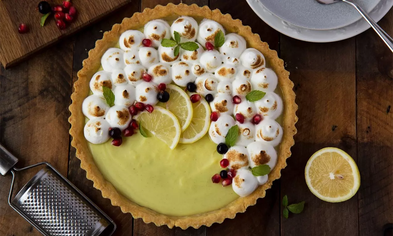 Lemongrass tart so svetložltou krémovou plnkou, ozdobený bielymi šľahačkovými kopčekmi, plátkami citróna, bobuľami a lístkami na drevenom stole