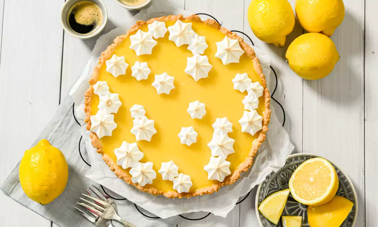 Leuchtend gelbe Zitronentarte mit goldbraunem Rand, dekoriert mit weißen Baiserhäubchen auf grauem Tuch, umgeben von frischen Zitronen und Besteck