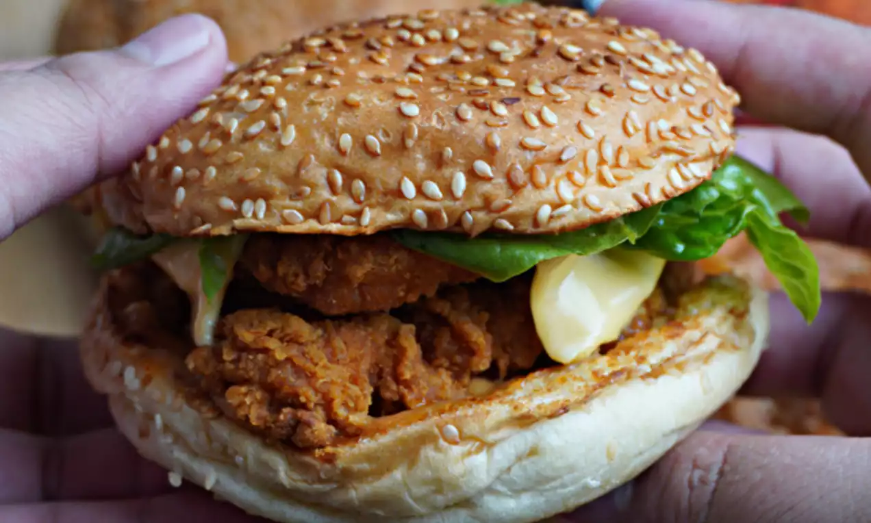 Sesame-seeded golden bun enclosing crispy orange-brown chicken, fresh green lettuce, and creamy pale yellow sauce in a close-up view