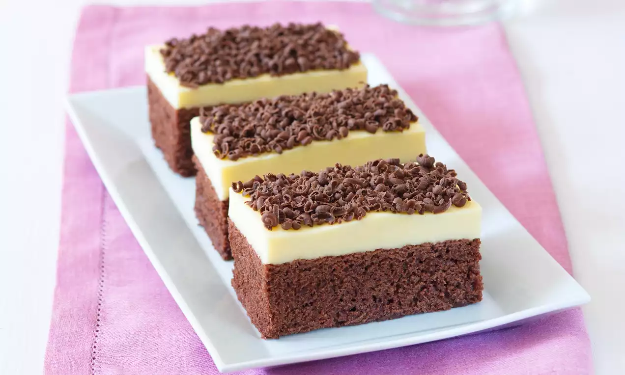 Tři hranaté kousky tmavého perníku s vrstvou světlého vanilkového krému, posypané drobnými čokoládovými hoblinkami, na bílém talíři položeném na růžovém ubrousku