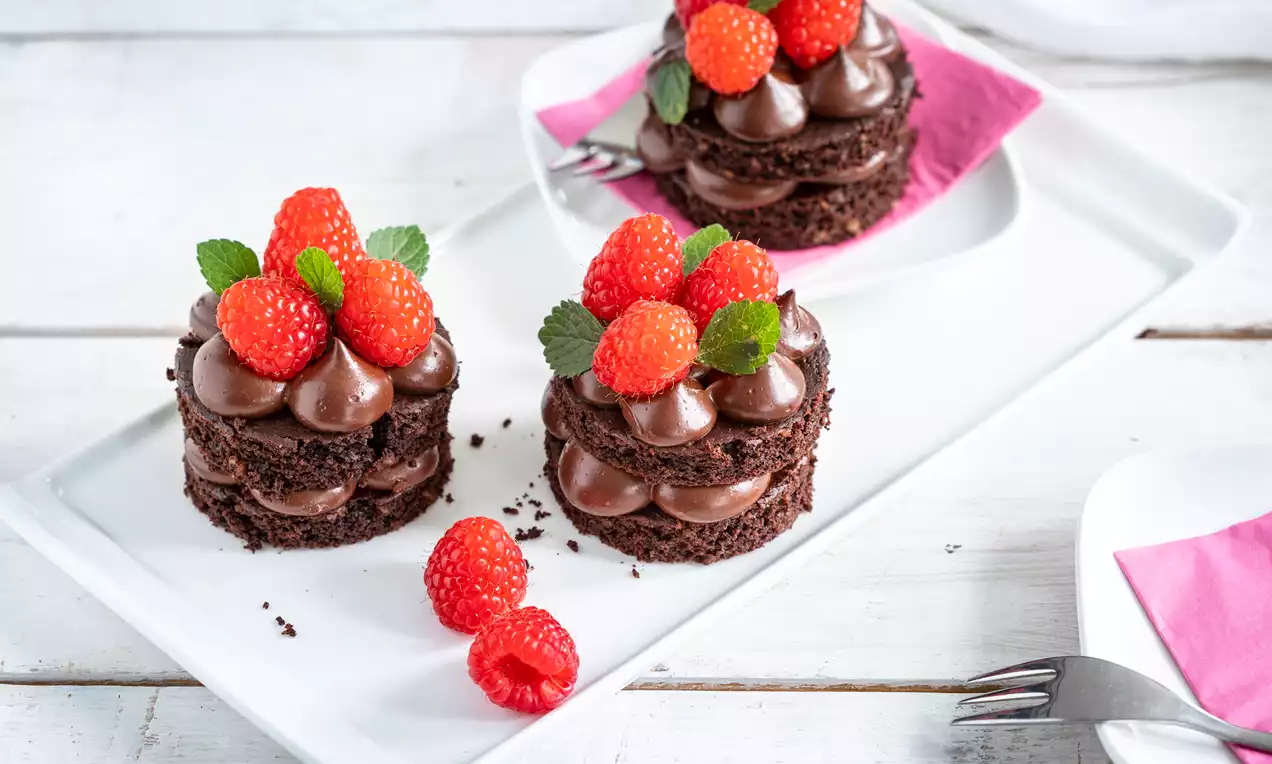 Zwei kleine vegane Schokoladenschichttorten mit glatter Creme, frischen Himbeeren und Minzblättern auf weißer Platte mit rosa Serviette