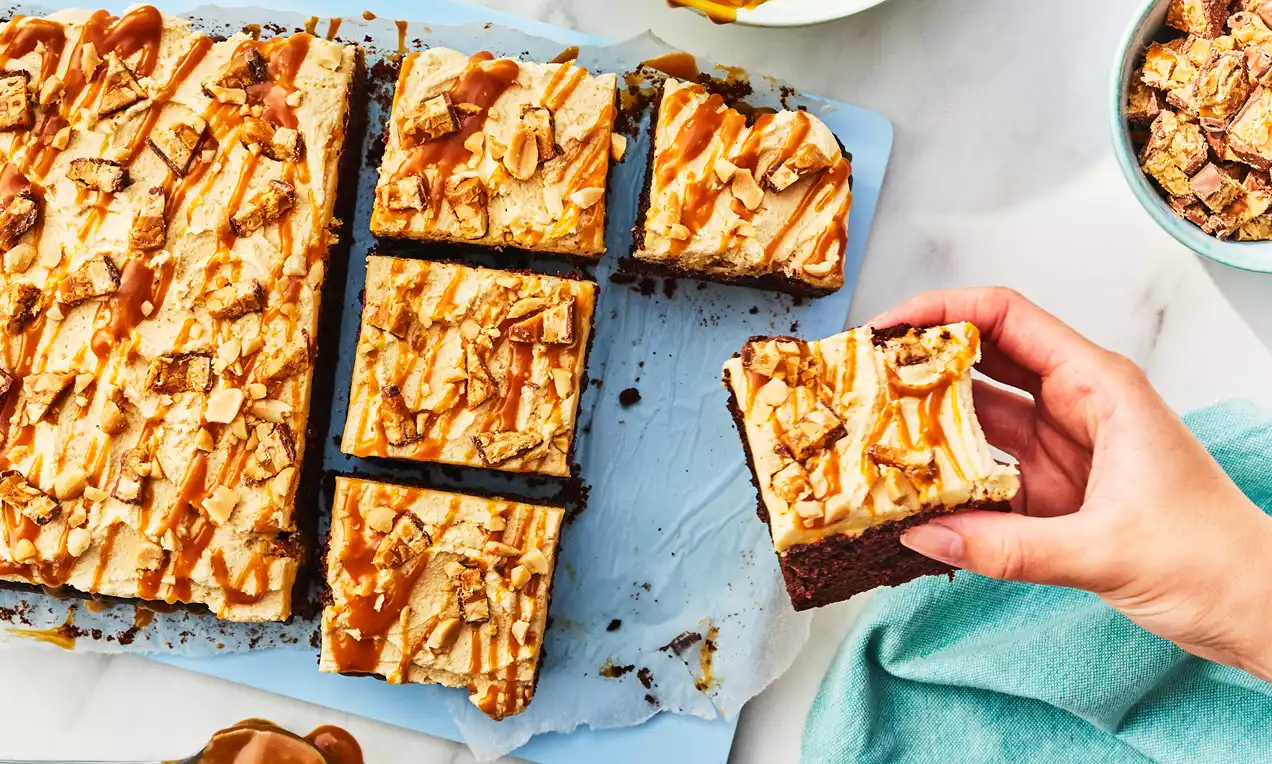 Squares of Snickers cake with a rich chocolate base, creamy topping, caramel drizzle, and chopped Snickers pieces on a blue tray