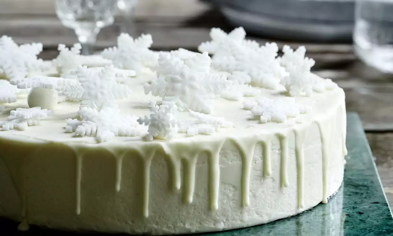 Rund kage dækket med lys glasur der drypper ned langs kanten, pyntet med hvide snefnug på et grønt fad