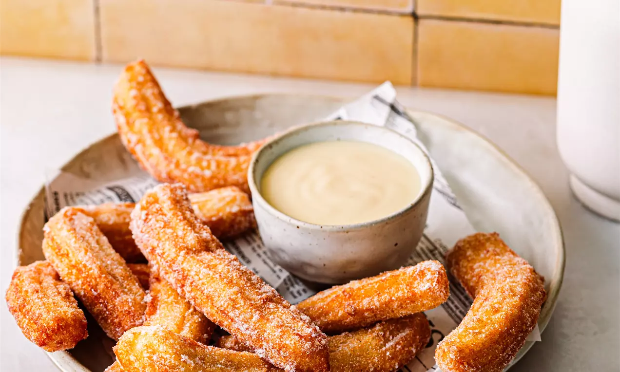 Sokerilla kuorrutetut, kullankeltaiset churrot aseteltuina metallivadin päälle, keskellä vaalea kastike metallikulhossa