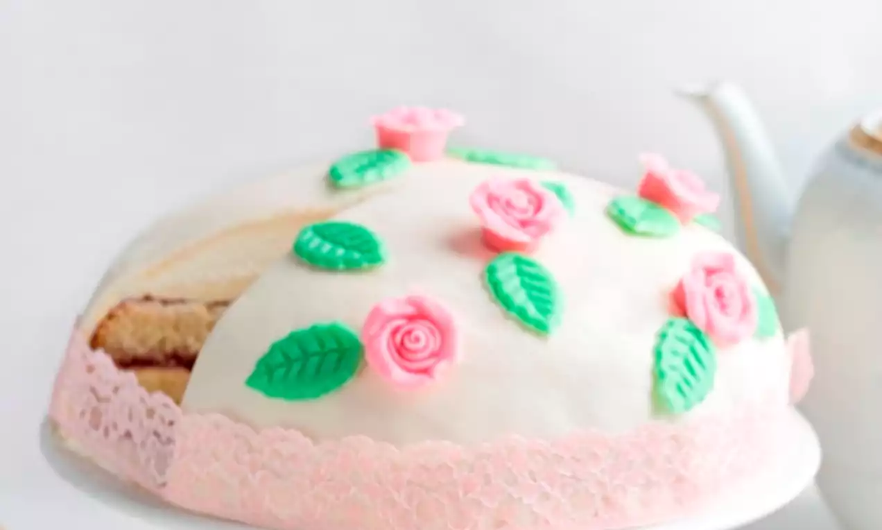 Vaaleanpunaisella pitsinauhalla koristeltu vaalea kupolimainen kakku, jonka pinnalla vaaleanpunaisia ruusuja ja vihreitä lehtiä valkoisella tarjoiluvadilla