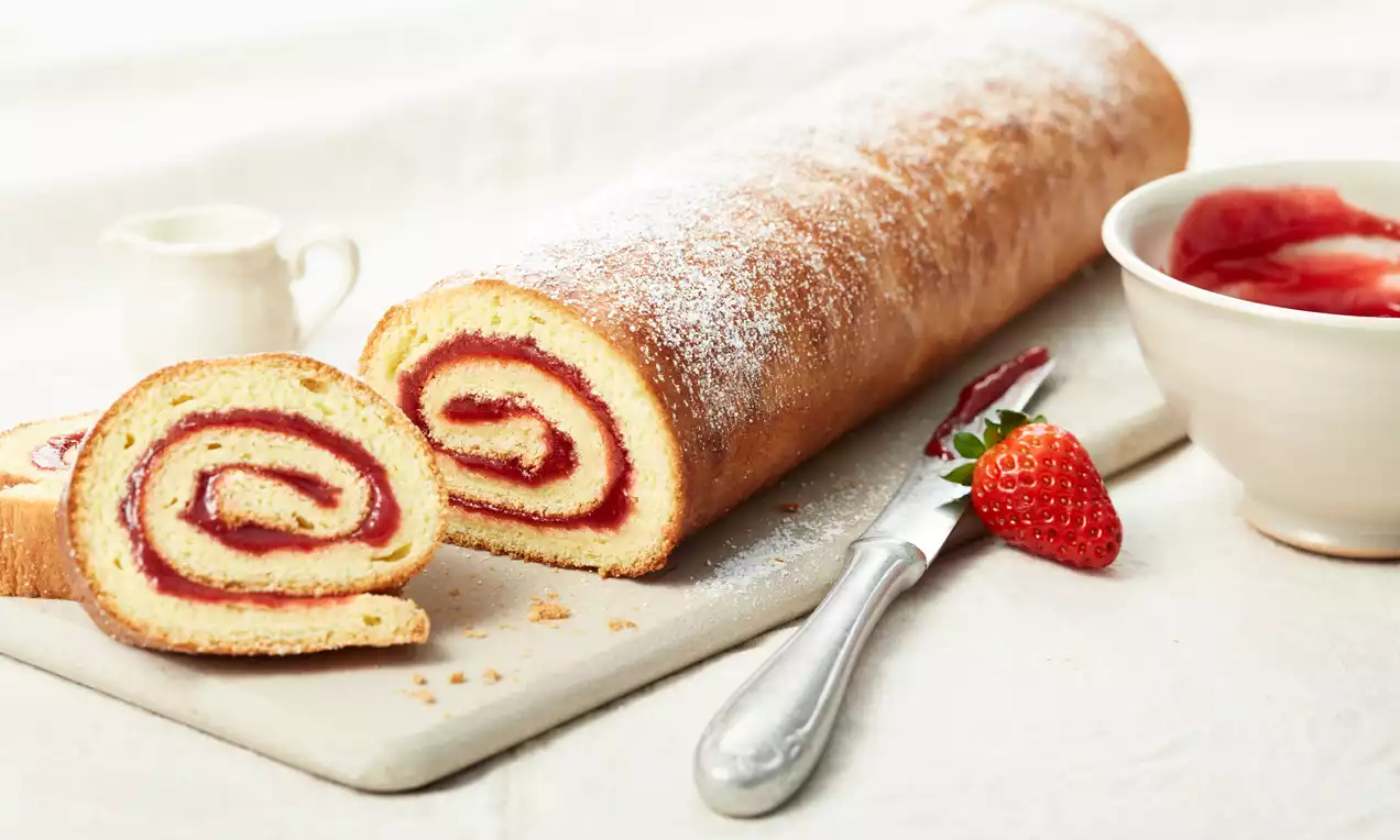 Goldbraune Biskuitrolle mit roter Fruchtfüllung, in Scheiben geschnitten auf heller Platte, daneben Erdbeeren, Messer und weiße Schale