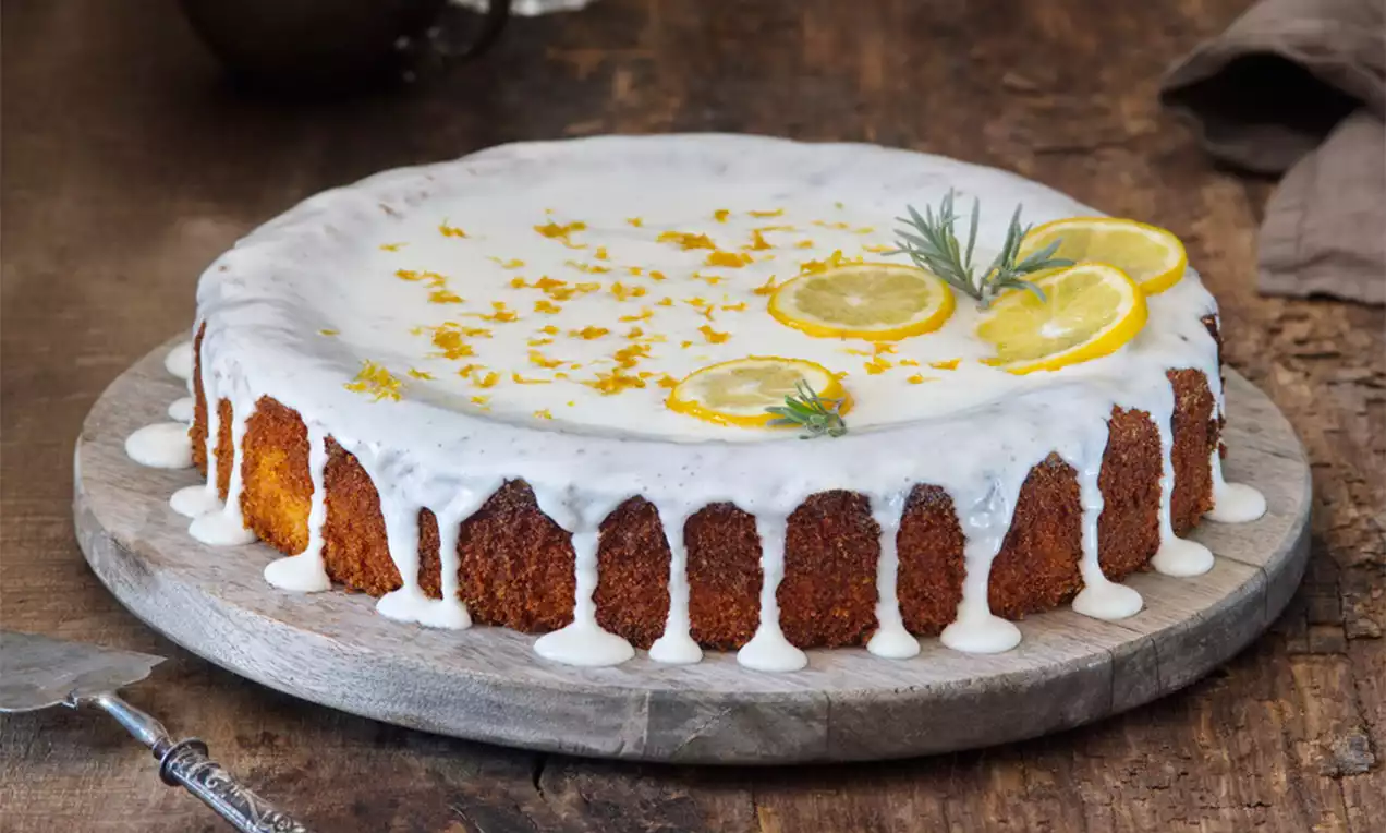 Polentový koláč s karamelovo-hnedým základom, poliaty bielou citrónovou polevou, ozdobený plátkami citróna a vetvičkou rozmarínu na drevenom podnose