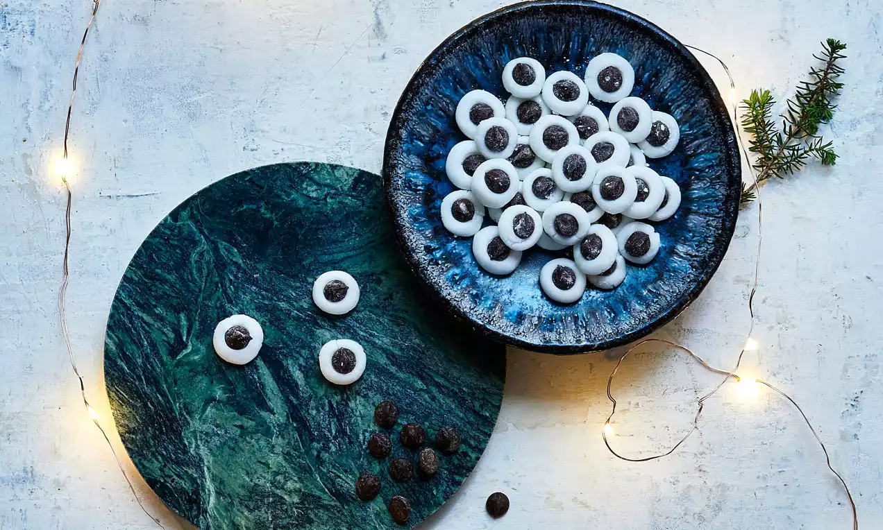 Hvide runde pebermyntepastiller med mørk chokoladeprik i midten på blå skål og grøn tallerken omgivet af små lyskæder og granpynt
