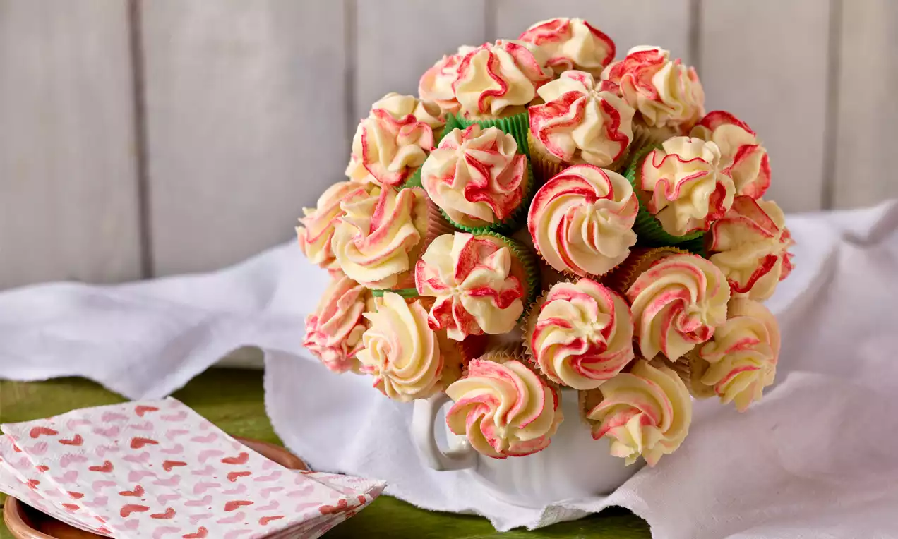 Ein Cupcake-Strauß aus kleinen, cremefarbenen und rosa glasierten Cupcakes in grünen Förmchen, arrangiert in einer weißen Teekanne