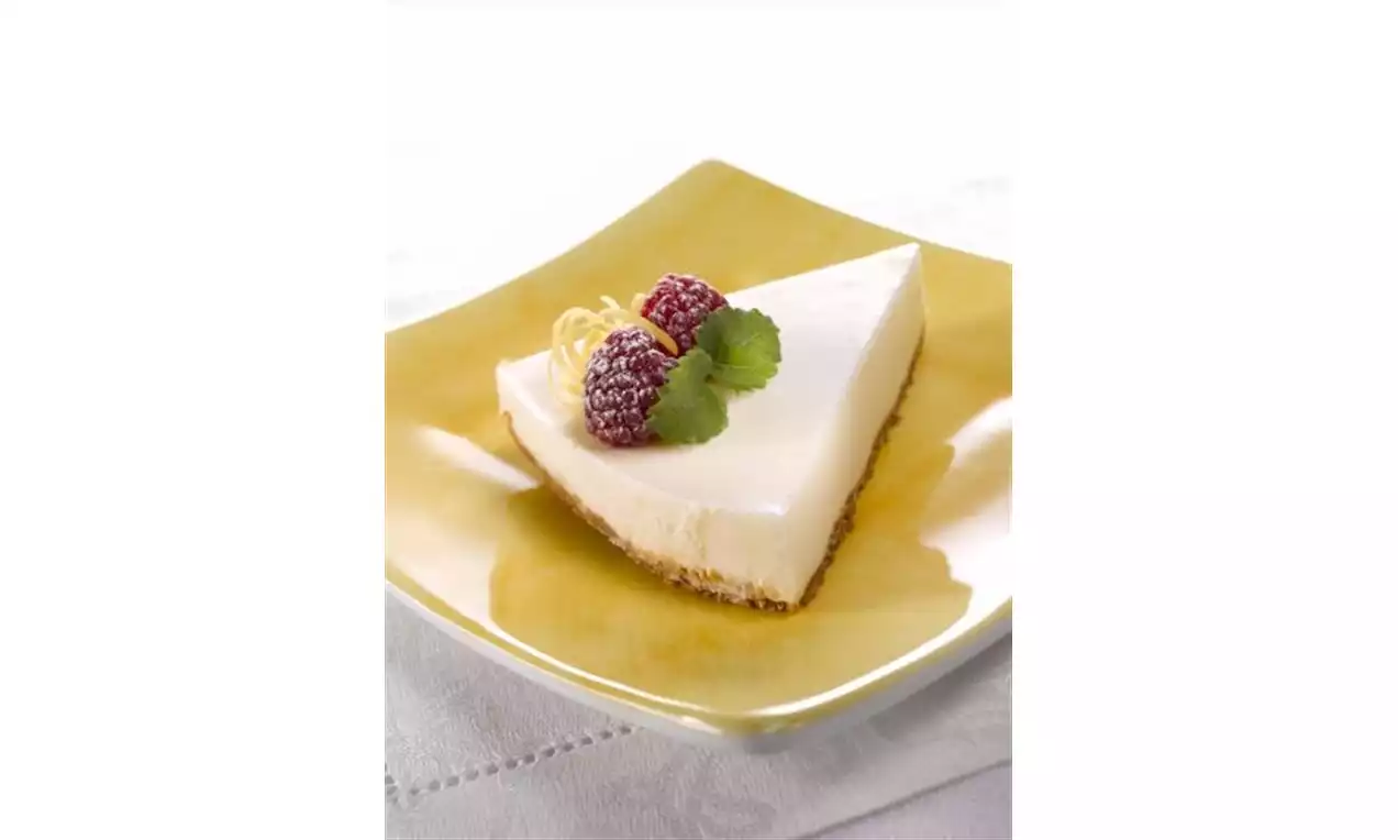 Citroncheesecake med ljus fyllning och gyllene kakbotten på gul tallrik, toppad med hallon, citronzest och gröna blad på vit duk