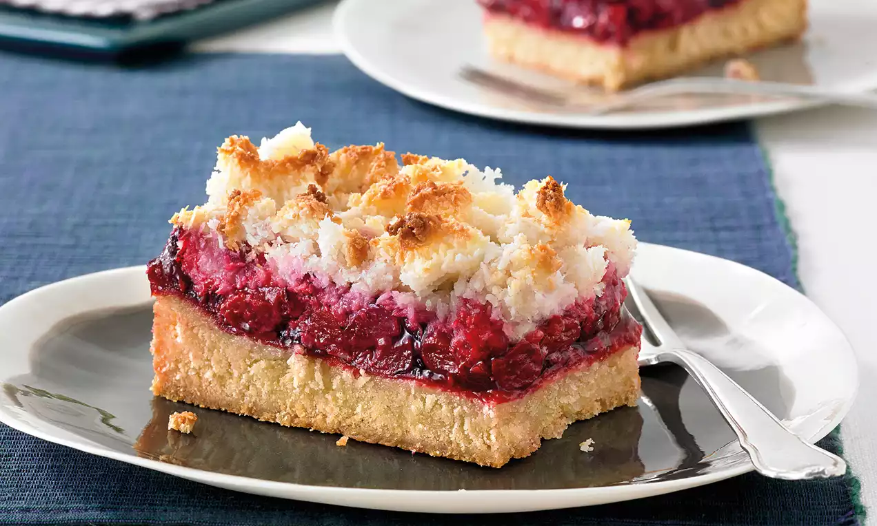 Ein Stück Kirsch-Kokos-Kuchen mit goldbraunem Butterboden, fruchtiger roter Kirschschicht und knuspriger Kokoshaube auf einem grauen Dessertteller