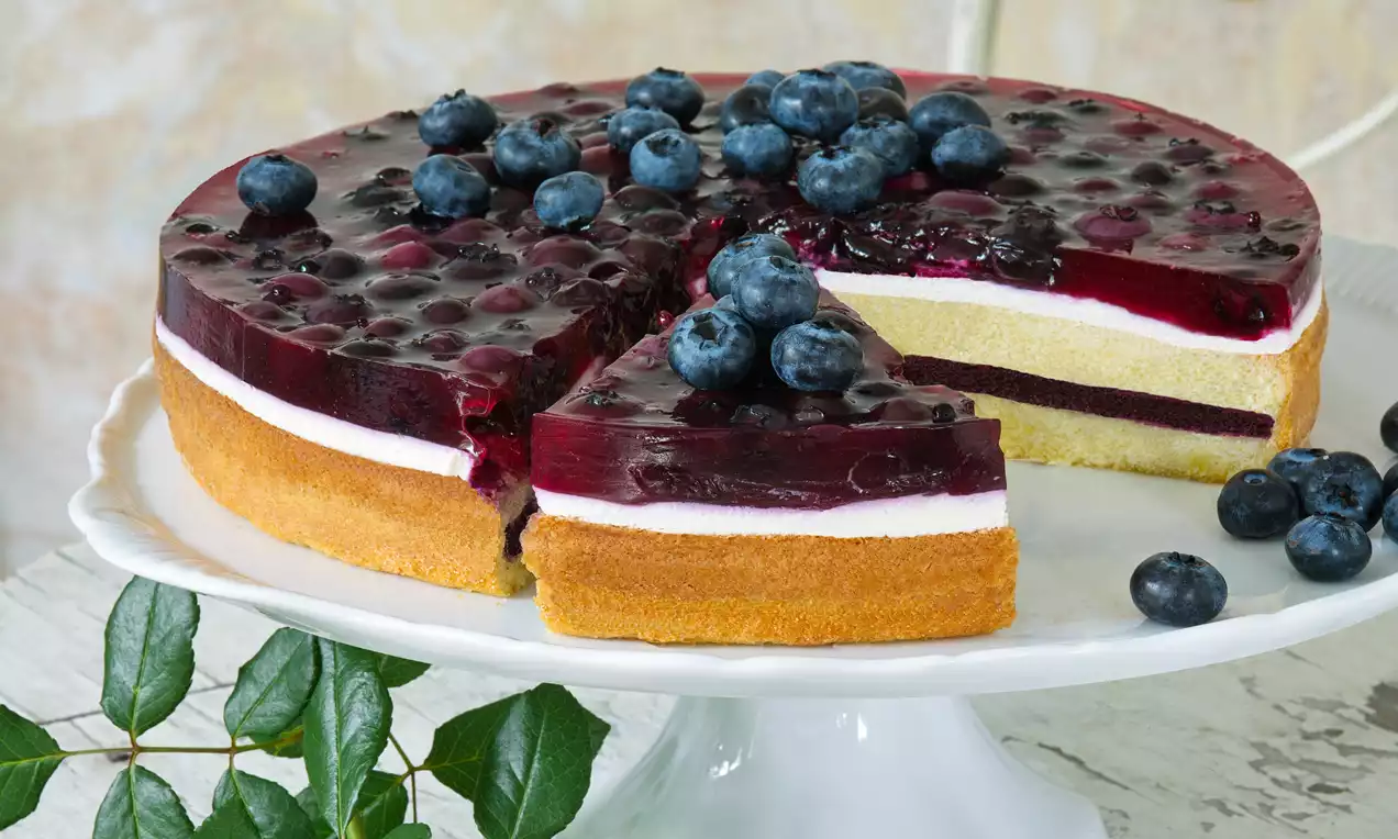 Borůvkový dort s piškótovým základom, vrstvou bieleho krému, želé bohato posiatym čučoriedkami, podávaný na bielom stojane s lístkami na stole