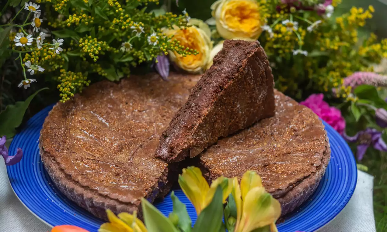 Čokoládová torta s bohatou tmavohnedou textúrou na modrom tanieri, obklopená pestrými kvetmi, s oddeleným trojuholníkovým rezom