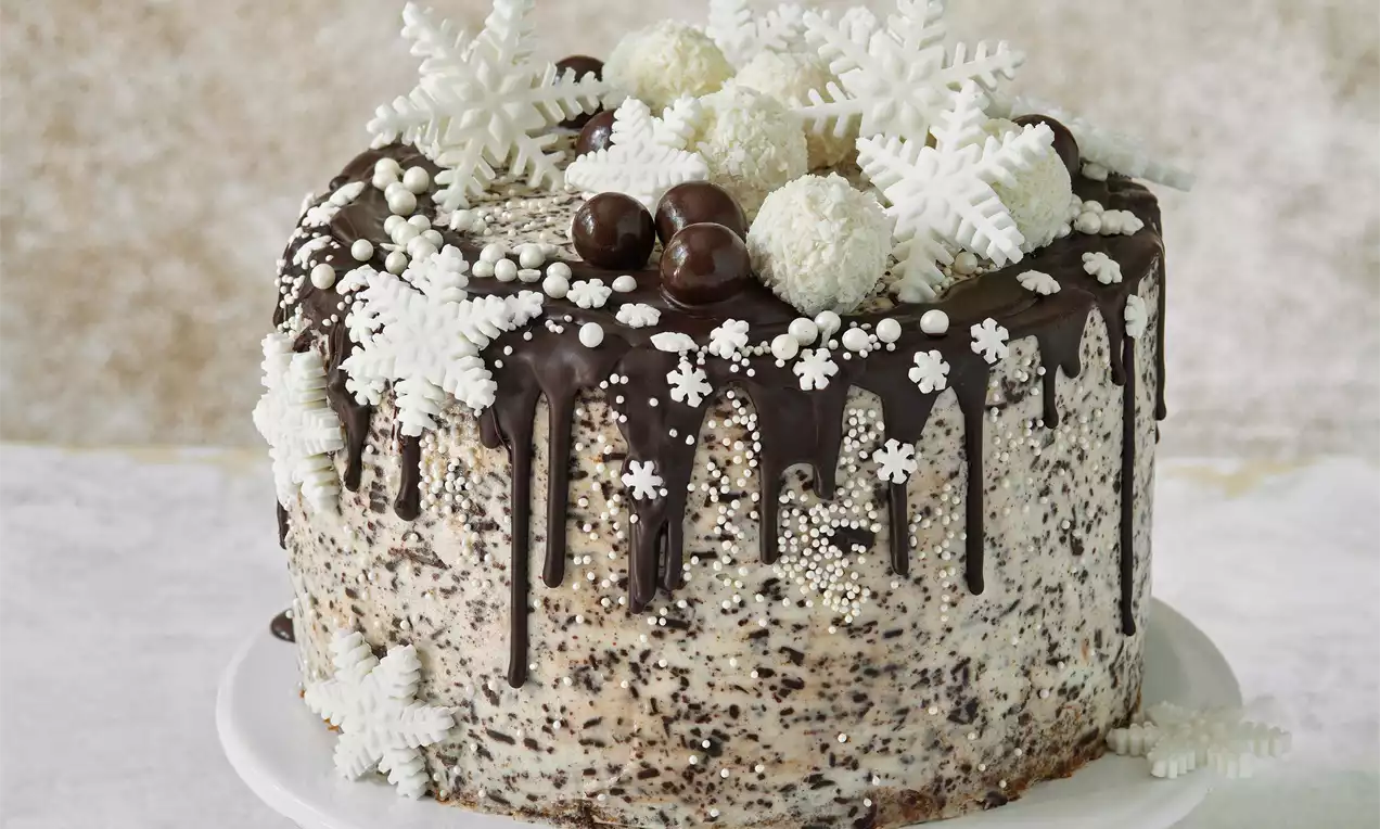 Zimný tort posypaný perleťovými vločkami s čokoládovou polevou, snehovými vločkami, bielymi guľkami a čokoládovými pralinkami na podnose