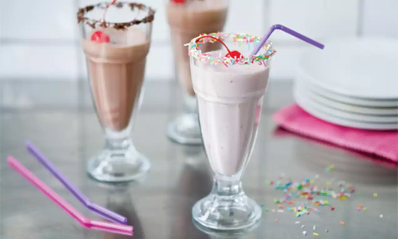 Vaaleanpunainen pirtelö pyöristettymuotoisessa lasissa värikkäillä strösseleillä ja kirsikalla koristeltuna, pöydällä pastellisävyisiä pillejä