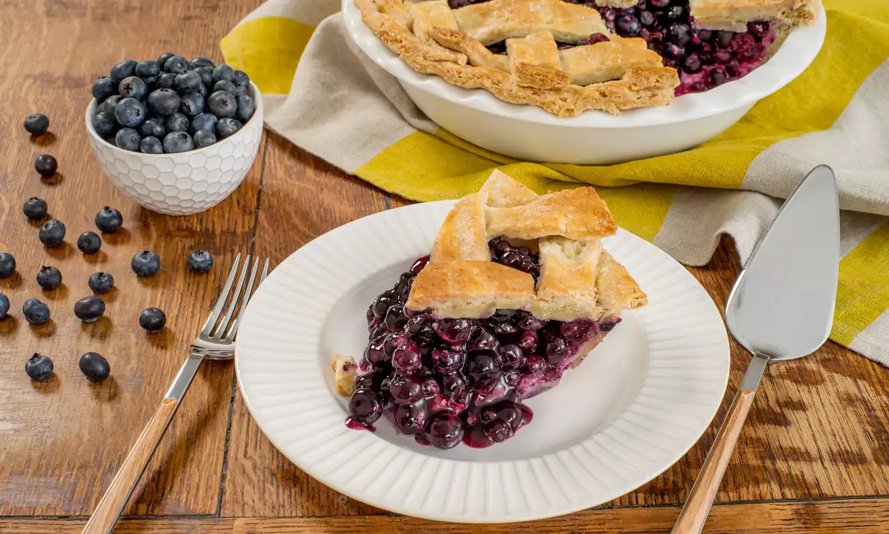 Tarte aux bleuets avec une croûte dorée en croisillons, garnie de fruits brillants et juteux, servie sur une assiette blanche avec une nappe jaune rayée