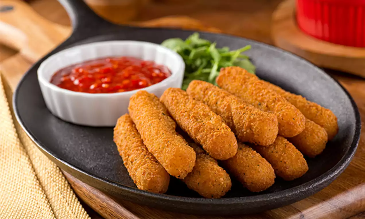 Golden, crispy mozzarella sticks arranged in a neat stack on a black skillet, served with red chili sauce in a white ramekin