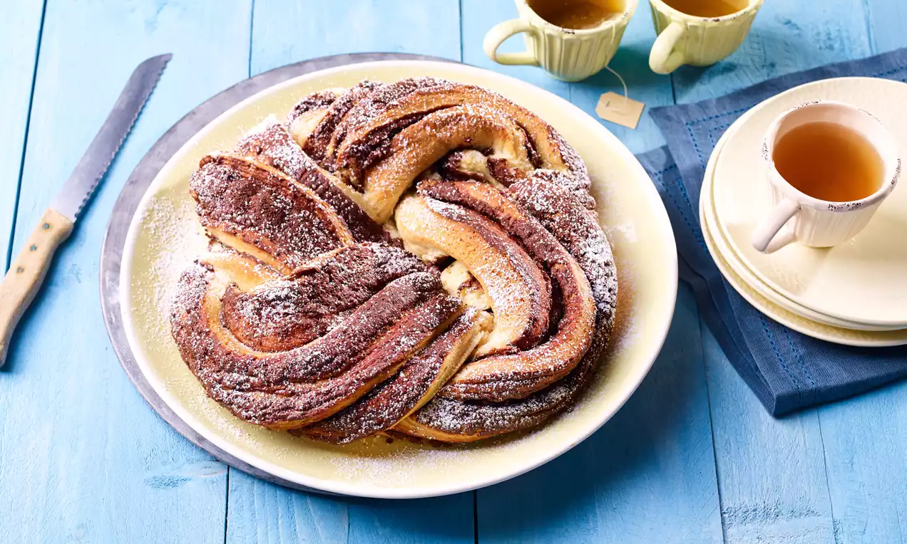 Gedrehter Schokoladenhefekranz mit goldbrauner Kruste, zartem Schokowirbel, leicht mit Puderzucker bestäubt auf weißem Teller