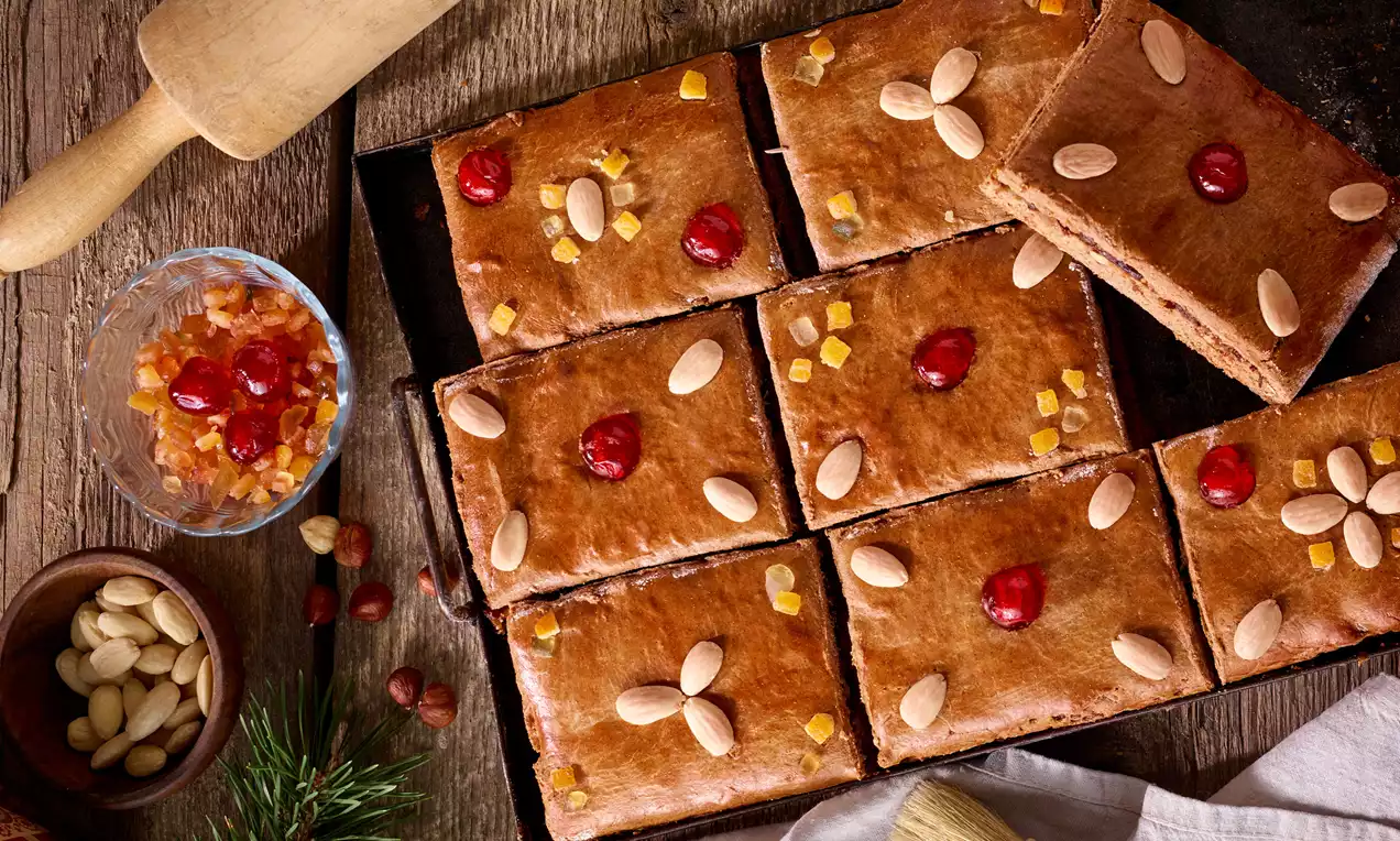 Tmavohnedé plátky perníka obdĺžnikového tvaru s lesklým povrchom, zdobené mandľami, červenými kandizovanými čerešňami a kúskami pomarančovej kôry