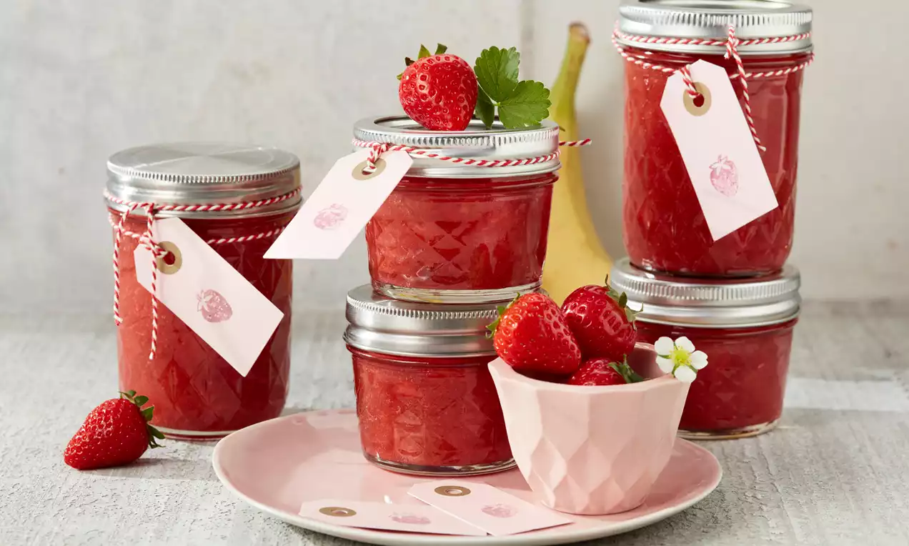 Gläser mit leuchtend roter Erdbeer-Bananen-Marmelade, teils mit Anhängern verziert, umgeben von frischen Erdbeeren und Banane, auf hellem Untergrund