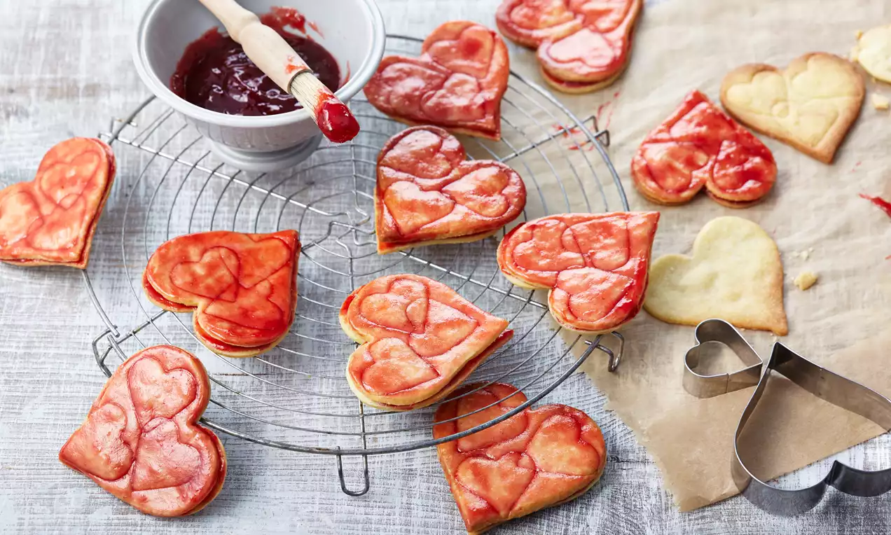 Szív alakú linzerek aranyló és málnaszínű mázzal, vékony mintázattal, rácson hűlve, egy tál piros lekvár és süteményszaggató mellett