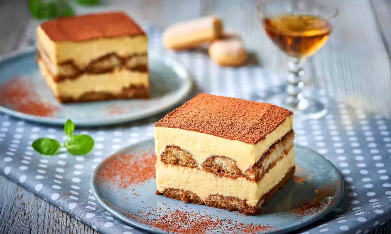 Kwadratowe kawałki tiramisu o kremowej, warstwowej strukturze z posypką z kakao na niebieskim talerzu z listkami mięty