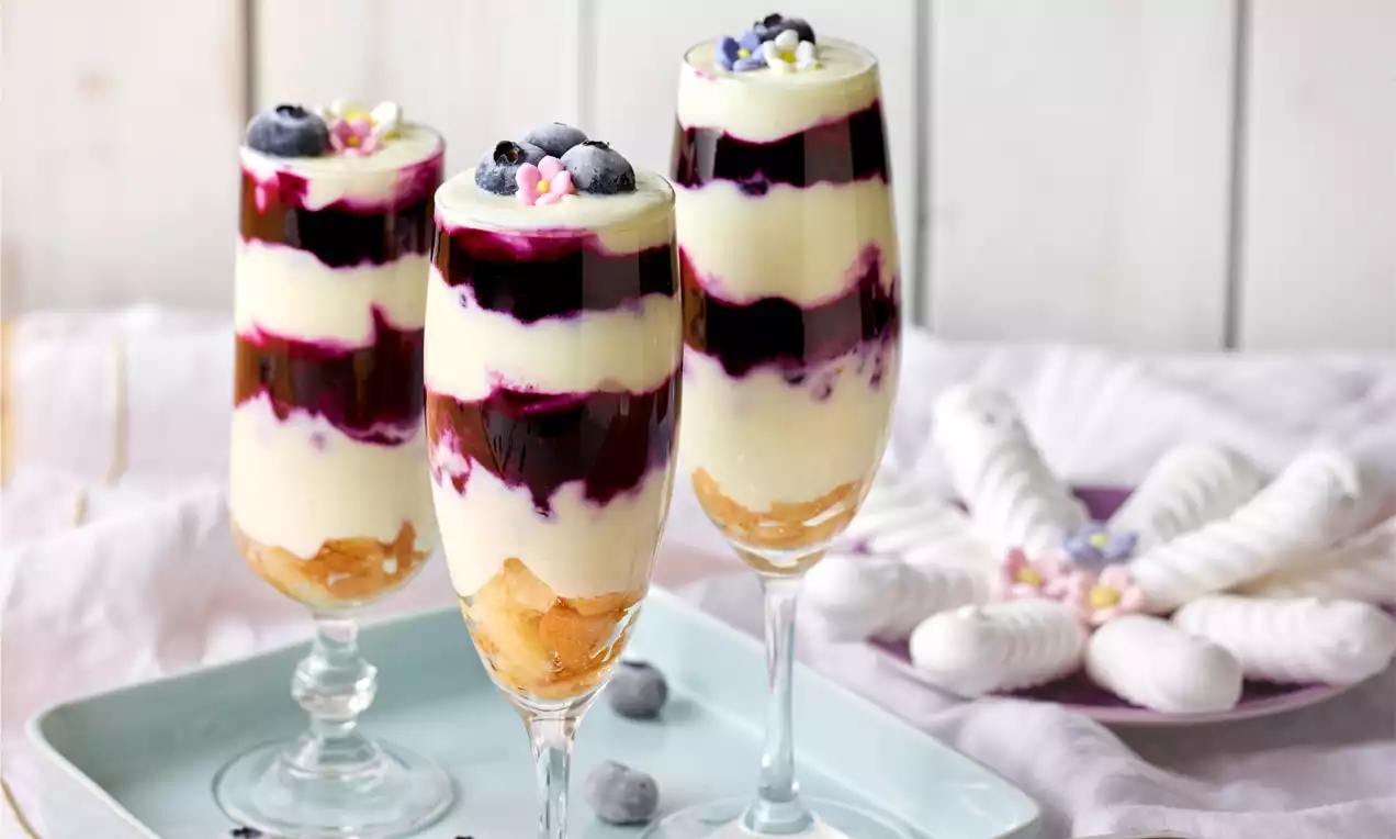 Schichtdessert in hohen Gläsern aus weißem Topfen und tiefvioletter Heidelbeersauce, dekoriert mit frischen Heidelbeeren und Zuckerstreuseln