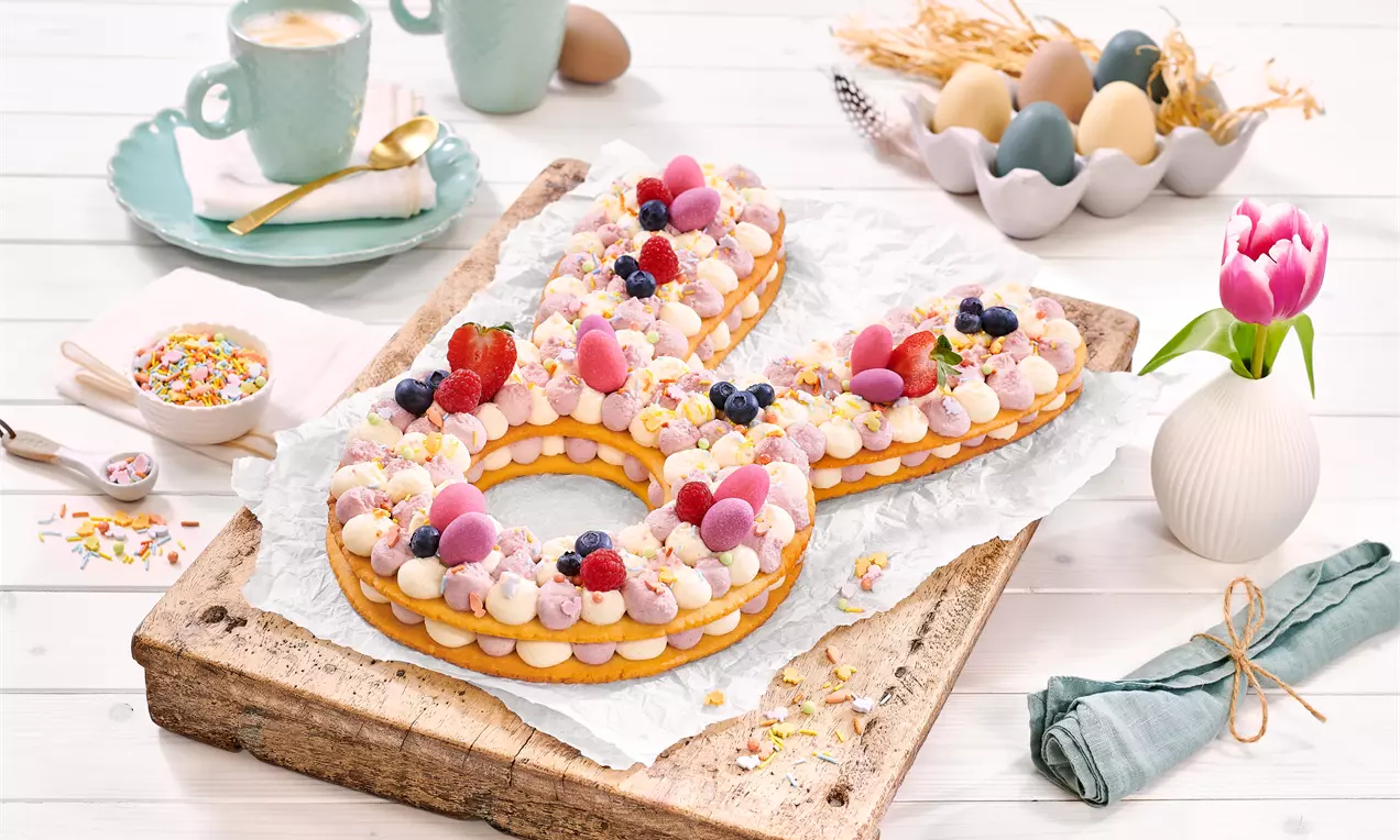 Veliko slovo oblikovano od dva sloja zlatnosmeđeg tijesta ukrašeno bijelim kremom, raznobojnim voćem i dekorativnim slatkišima na drvenoj podlozi