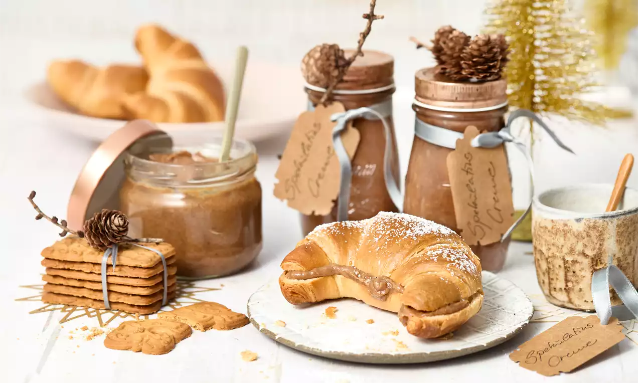 Goldbraunes Croissant mit cremiger Spekulatiusfüllung auf einer rustikalen Platte, umgeben von Gläsern, Keksen und weihnachtlicher Dekoration