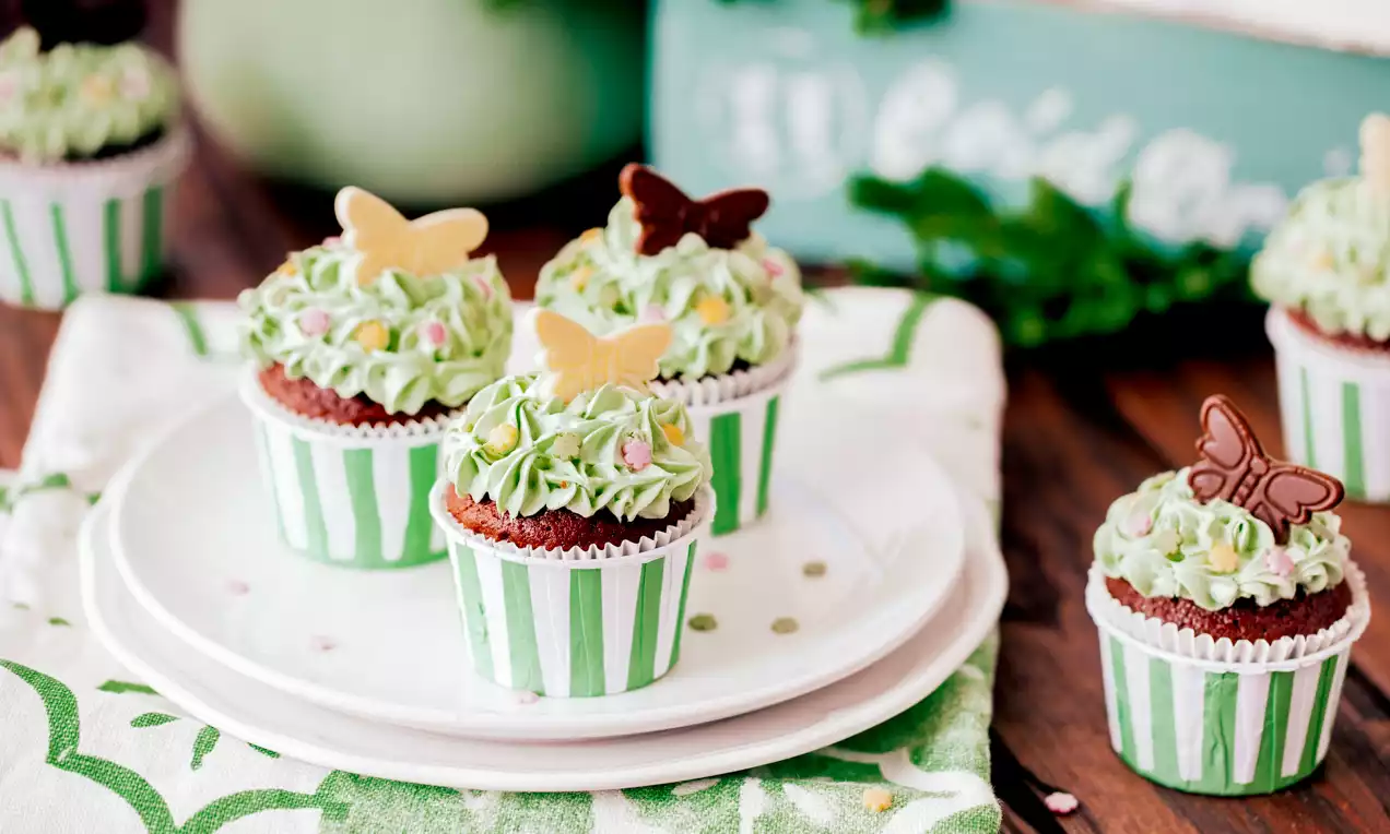 Cupcakes mit grün-weiß gestreiften Papierförmchen, hellgrünem Sahnehäubchen, bunten Perlen und Schmetterlingsdekoration auf weißem Teller