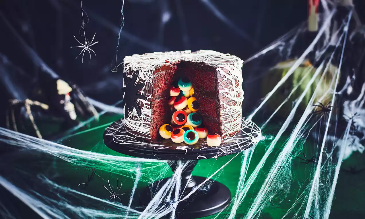 Chocolate cake with a glossy dark surface wrapped in white web-like icing, revealing a hollow filled with vibrant candy eyeballs on a dark stand
