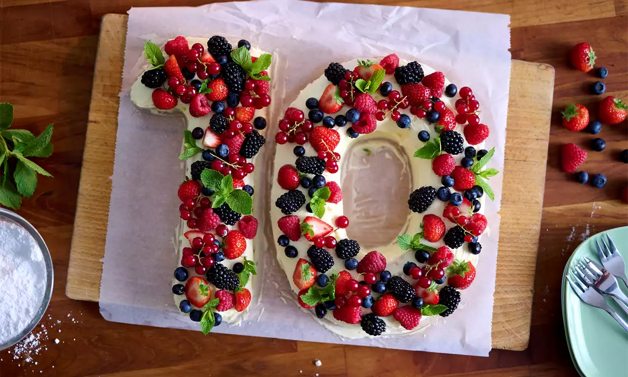 Cijfercake gevormd als 10, met witte crème, rijkelijk versierd met verse rode en blauwe bessen, muntblaadjes op bakpapier en houten plank