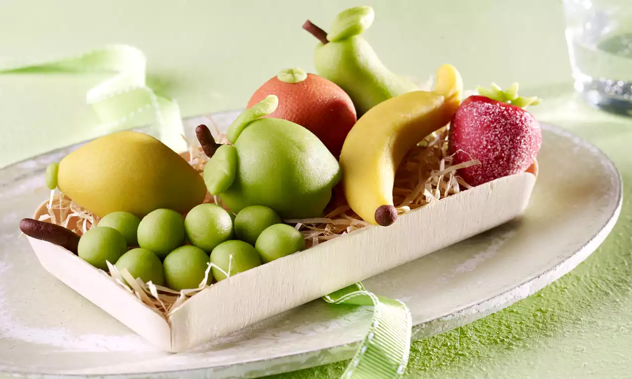 Kolorowe marcepanowe owoce o gładkiej powierzchni w drewnianej tacce na sianku, udekorowane zieloną wstążką na pastelowym tle