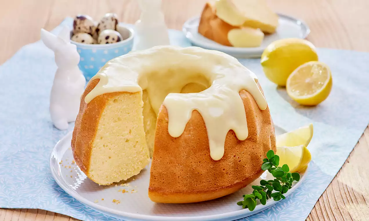 Babka jogurtowa w kształcie pierścienia o złocistym kolorze z cytrynowym lukrem, na białym talerzu z plasterkami cytryny i gałązką zieleniny