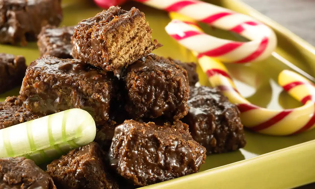 Cubetti di pane speziato scuro con glassa brillante disposti su un piatto verde, accanto a bastoncini di zucchero colorati