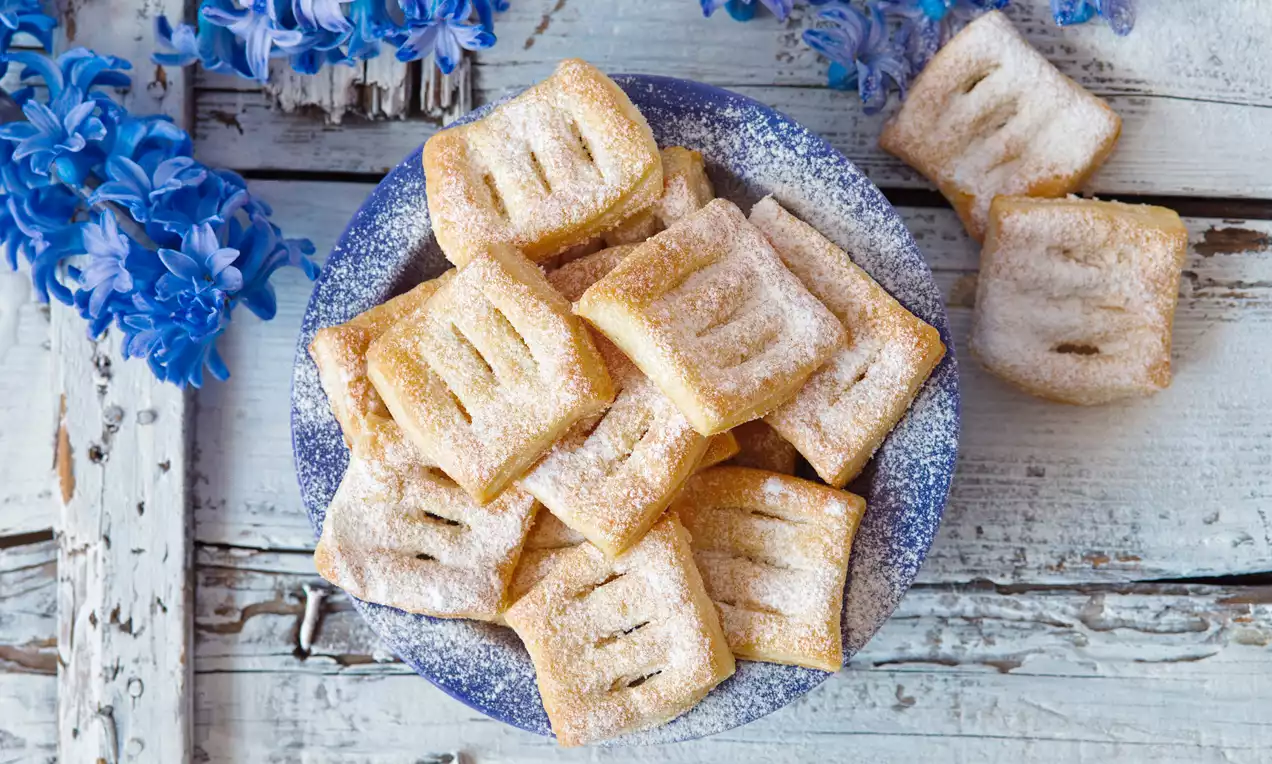 Zlatavé čtvercové Boží milosti s jemnými vroubky, posypané moučkovým cukrem, na modrém talíři vedle čerstvých modrých květů hyacintu