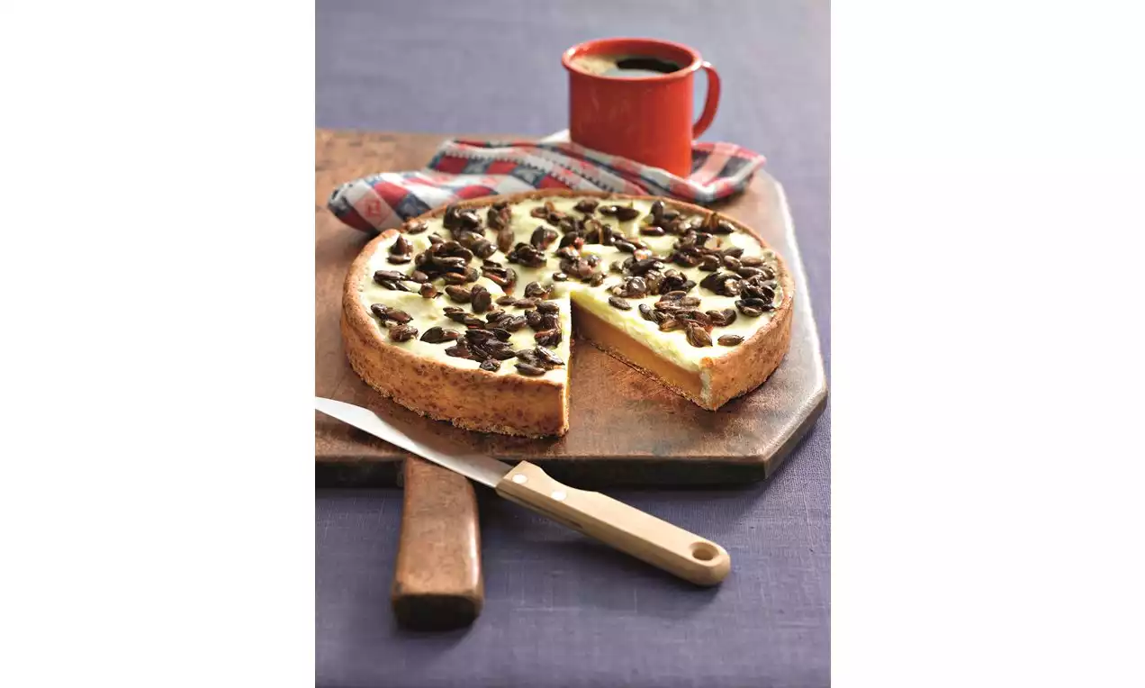 Dýňový koláč na dřevěné desce s hladkým krémem světle oranžové barvy, posypaný praženými semínky, vedle lžíce, hrníčku a látky