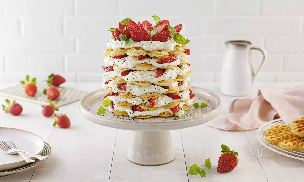 Ein gestapelte Waffelkuchen mit Schichten aus hellem Teig, weißer Creme, roten Erdbeeren und grüner Minze auf einer eleganten Kuchenplatte
