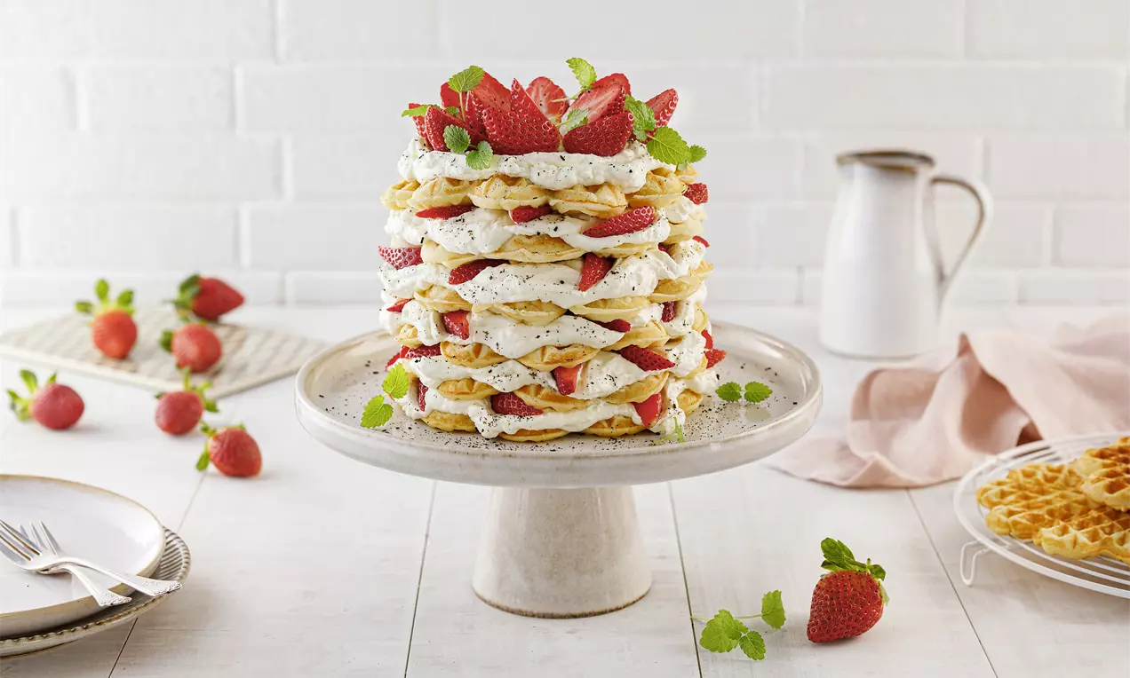 Hohe Torte aus mehreren Lagen goldgelber Waffeln, weißer Sahnecreme mit Vanille und roten Erdbeeren, garniert mit Minzblättern auf Tortenplatte