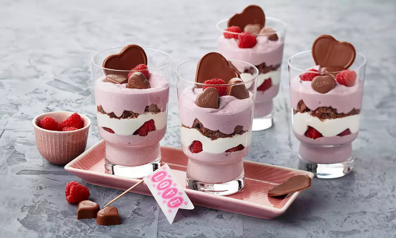 Schichtdessert in Rosa und Weiß mit frischen Himbeeren, Schokostückchen und Herzkeksen, serviert in Glasbechern auf rosa Keramikplatte