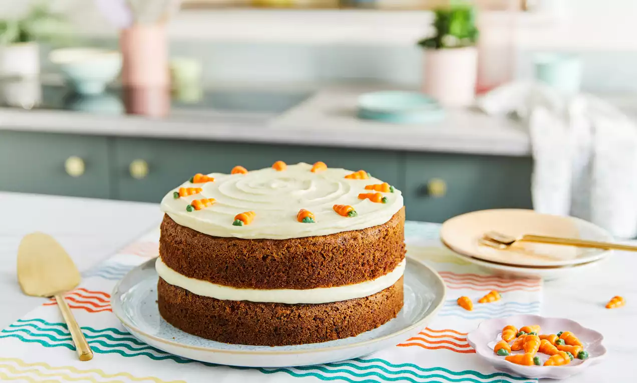 Carrot cake with two layers of rich brown sponge, smooth white frosting swirls, and tiny orange and green icing carrots on a white plate
